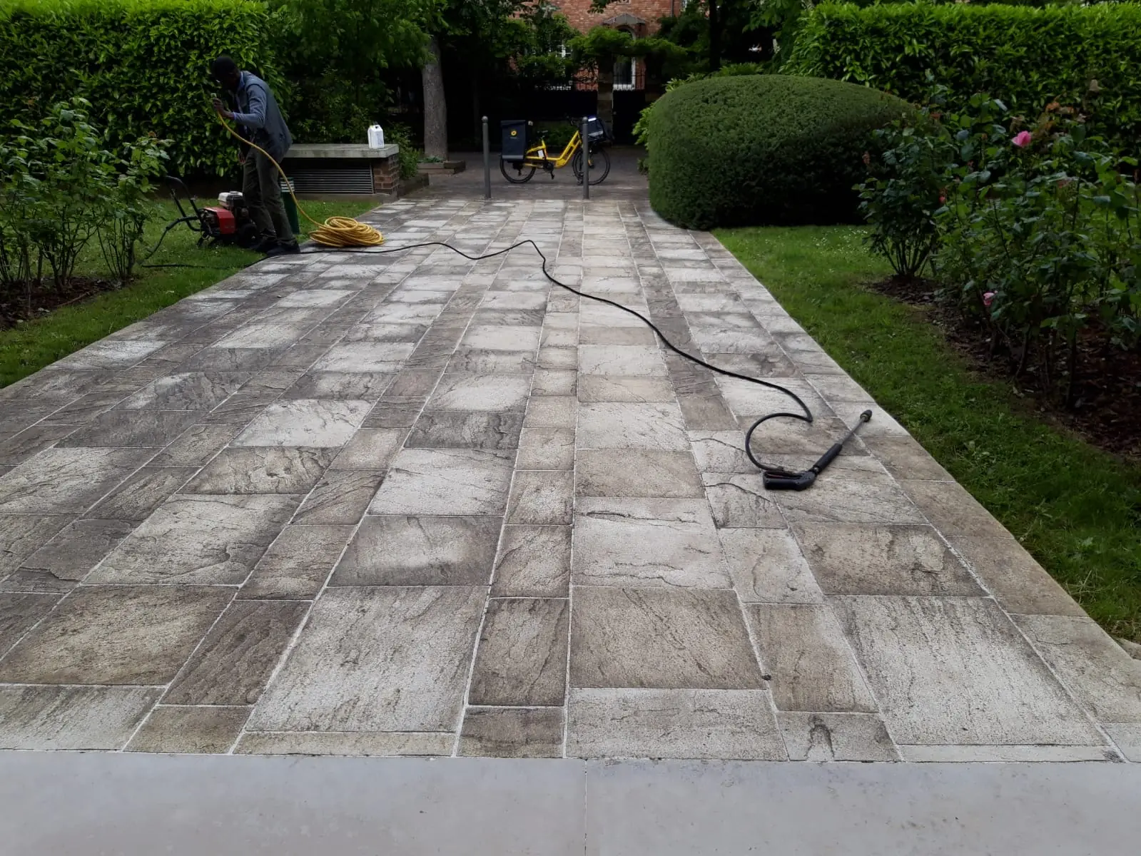 Allée de jardin en dalles en cours de nettoyage, le nettoyeur haute pression au fond entre les haies