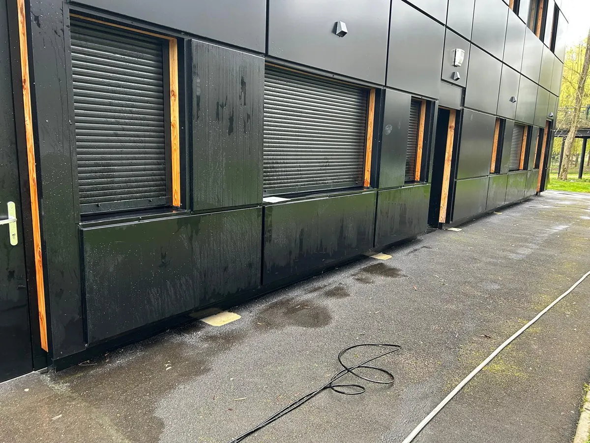 Façade en bardage noir d’un bâtiment, volets roulants et encadrements en bois, le pied de façade et le trottoir encore mouillés après le lavage