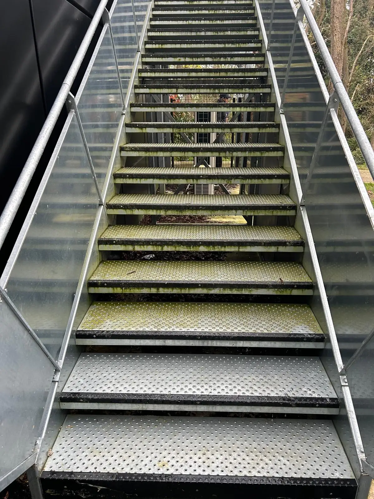 Escalier extérieur métallique vu d’en bas, avant le nettoyage : marches en tôle striée verdies de mousse et de dépôts