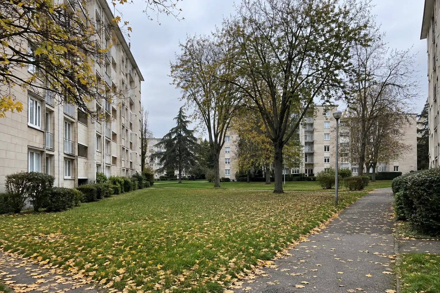 Barres de logements à parement de pierre claire, pelouses et grands arbres d’automne, allée piétonne entre les immeubles